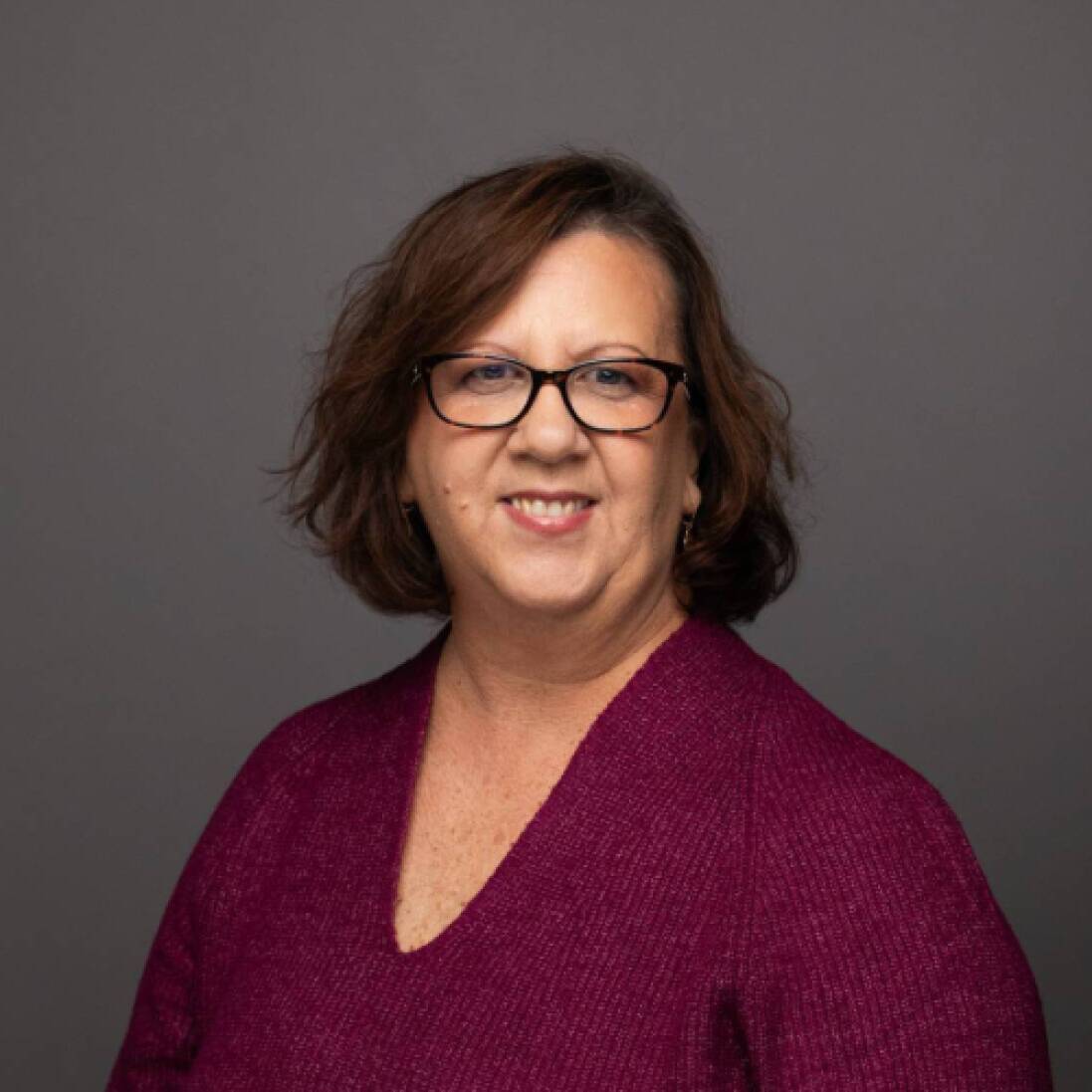 A woman with short brown hair and brown glasses wearing a burgandy long sleeve blouse smiles at the camera in front of a gray studio background